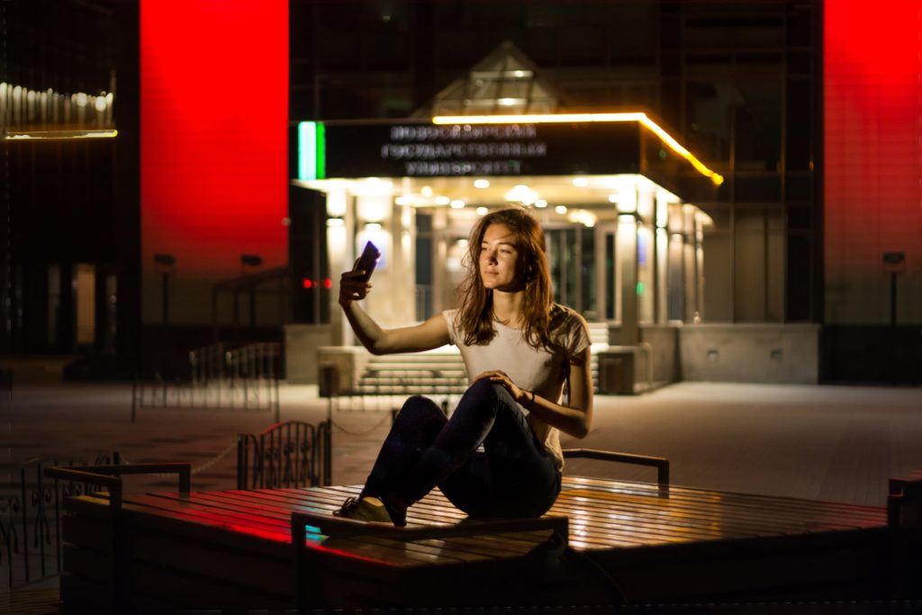 a girl sitting on a bench and taking a selfie, night, lights, red walls, alla biriuchkova